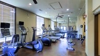 This fitness center is designed to support a well-rounded exercise routine within a single-floor layout. The space is outfitted with a variety of cardio equipment, including multiple treadmills, elliptical trainers, and stationary bikes from brands such as Life Fitness and True. A dedicated free weight area lines one wall, featuring a fully stocked dumbbell rack with weights arranged in ascending order for convenient access.

Large windows fitted with horizontal blinds allow natural light to fill the room, contributing to an open and energizing atmosphere. A full-length mirror spans one wall, visually expanding the space and allowing users to monitor their form during workouts. Ceiling fans provide additional air circulation throughout the room, complementing the overhead HVAC system.

The flooring is a durable, dark-toned surface well-suited for high-traffic fitness use. Recessed lighting supplements the natural light, ensuring the space remains well-illuminated at all times. French doors visible in the mirrored reflection suggest access to an adjacent area or outdoor space, adding to the overall sense of openness.