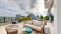 Located in Fort Lauderdale, FL, this rooftop terrace offers an elevated outdoor living experience with sweeping panoramic views of the city skyline and surrounding landscape. The space is defined by light-toned porcelain tile flooring that extends seamlessly from the interior lounge to the open-air deck, creating a cohesive and sophisticated aesthetic throughout.

The outdoor terrace is generously proportioned and thoughtfully arranged with multiple seating areas, including a large sectional sofa paired with a coffee table, as well as accent chairs in natural rattan and navy blue — providing both casual lounging and conversational settings. Raised planter beds filled with ornamental grasses and tropical greenery serve as architectural focal points, adding a vibrant natural element against the urban backdrop.

The interior lounge connects directly to the terrace through full-height glass walls and floor-to-ceiling glass doors, allowing abundant natural light to flood the indoor space while framing dramatic sky and cityscape views. Inside, rattan chairs, colorful side tables, and additional soft seating create a relaxed, resort-inspired atmosphere.

The glass perimeter railing along the terrace preserves unobstructed sightlines across the Fort Lauderdale skyline, making this rooftop an ideal space for both social gatherings and quiet enjoyment of the South Florida skyline.