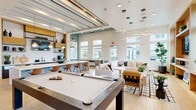 Located in Murrieta, CA, this single-floor community lounge offers a thoughtfully designed open-plan layout that seamlessly integrates a kitchen and living area. The space is defined by its contemporary architectural details, including a striking slatted ceiling with exposed beams accented in warm wood tones and dark metal, complemented by light hardwood-style flooring throughout.

The kitchen area centers around a large, stone-topped island with seating for multiple guests, illuminated by modern globe pendant lights. Dark cabinetry lines the back wall, flanked by open wood and metal shelving that adds warmth and visual interest. A mounted flat-screen TV and a coffee station complete this functional yet stylish zone.

The living area transitions naturally from the kitchen, featuring a comfortable sectional sofa, patterned area rug, and a built-in wood shelving unit with a second TV. A statement starburst chandelier anchors the seating area, while oversized windows and transom windows along the perimeter flood the room with natural light and provide views of the surrounding community.

A full-size billiards table occupies the central gathering space, making it ideal for recreation and social interaction. Large planters with lush greenery add life and color throughout, reinforcing the space's welcoming, resort-inspired ambiance.