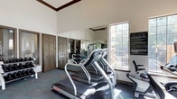 This fitness center is a spacious, single-floor facility designed to support a comprehensive workout experience. The room is defined by its impressively high ceilings, which create an open, airy atmosphere and contribute to a sense of generous volume throughout the space. Large windows, including clerestory-style windows near the roofline, flood the interior with abundant natural light, while glass double doors at one end provide direct outdoor access and additional brightness.

Floor-to-ceiling mirrors line the walls, visually expanding the space and allowing users to monitor their form during exercise. The facility is well-stocked with a wide range of cardio equipment, including treadmills, elliptical trainers, upright and recumbent stationary bikes, and a rowing machine. A dedicated strength training area features a multi-station cable machine, a weight bench, and a full rack of dumbbells, along with exercise balls for functional training.

The flooring consists of a durable, teal-toned carpet suited for fitness use. Overhead ceiling fans provide air circulation throughout the workout area, complementing the climate control systems. Crown molding details and warm-toned door frames add a refined architectural touch to the otherwise functional space. The overall ambiance is bright, clean, and motivating, making it a well-rounded facility for a variety of fitness routines.