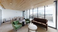 This single-floor amenity lounge is designed to offer a refined yet comfortable gathering space with a distinctly modern aesthetic. The most striking architectural feature is the warm, light-toned wood-planked ceiling, which spans the entire space and adds natural texture and warmth, contrasting beautifully against the crisp white walls and neutral-toned carpet flooring.

Floor-to-ceiling glass walls and sliding doors line the exterior-facing side of the space, flooding the interior with abundant natural light while offering sweeping views of a city skyline. These expansive windows connect the interior seamlessly to an adjoining outdoor terrace, which is furnished with high-top tables and bar-height seating—ideal for enjoying the elevated vantage point.

Inside, the lounge is organized into distinct functional zones. A seating area anchors one side of the room, furnished with a tufted dark-brown sofa, a round low coffee table with a wire base, and a pair of plush green velvet accent chairs around a small side table. A wall-mounted flat-screen television with a soundbar is positioned across from the sofa, making the space well-suited for casual viewing.

Adjacent to the lounge area sits a dining space with a rectangular wood table and coordinating ladder-back chairs, comfortably seating six. Multiple doorways at the rear of the space provide access to additional rooms and corridors, enhancing the overall functionality of the layout.