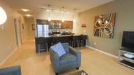 This single-floor space showcases an open-concept layout that integrates the kitchen and living area into one cohesive, functional environment. The kitchen is outfitted with rich dark cabinetry contrasted by warm wood-toned upper cabinets, stainless steel appliances including a full-size refrigerator and range with microwave, and a dark countertop that extends into a breakfast bar. The bar seating area accommodates multiple stools, making it an ideal spot for casual dining or entertaining. Pendant lighting hangs above the bar, complementing a track lighting system overhead that provides adjustable illumination throughout the space. The living area flows naturally from the kitchen, anchored by a plush gray armchair accented with a patterned blue pillow. Light hardwood-style flooring runs throughout, enhancing the sense of openness and continuity. Neutral warm-toned walls serve as a backdrop for colorful decorative artwork, adding personality to the space. The overall ambiance is modern and casual, balancing practical kitchen functionality with a comfortable, inviting lounge atmosphere.