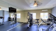 This single-floor fitness room is thoughtfully designed to support a well-rounded workout routine. The space is outfitted with a multi-station cable strength machine positioned at the center, offering a variety of resistance-based exercises, while a cardio machine — likely a treadmill or incline trainer — occupies the foreground. Dark rubber flooring covers the entire room, providing both safety and durability underfoot.

A full-length mirror spans one wall, visually expanding the space and allowing users to monitor their form during exercises. A flat-screen television is mounted adjacent to the mirror, adding an entertainment option for longer workout sessions. Multiple windows along the walls invite natural light into the room, creating a bright and energizing atmosphere, while a wall-mounted air conditioning unit ensures comfort during use.

The ceiling features recessed lighting alongside a ceiling fan with an integrated light fixture, maintaining airflow and adequate illumination throughout the room. The warm, neutral-toned walls contribute to a clean and focused environment, making this fitness room a functional and inviting space for daily exercise.