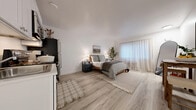 Located at 8835 S Hosmer St in Tacoma, WA, this efficiently designed one-bedroom, two-bathroom unit makes excellent use of its compact footprint across a single floor. The spacious primary bedroom serves as the central living area, featuring light wood-style flooring, crisp white walls, and a large window dressed with sheer curtains that allow abundant natural light to fill the room. A built-in kitchenette along one wall includes white shaker-style cabinetry, a stainless steel refrigerator, range, and microwave, providing all essential cooking amenities within easy reach. The room also accommodates a dining area with a rustic wooden table and stools, along with ample space for a full-sized bed and additional furnishings. The bathroom area is divided into two functional spaces: a shared vanity zone with a white shaker-style cabinet, a large mirror, and a textured accent wall, plus a separate water closet with light tile flooring and a full bathtub with a tiled surround. A hallway connects the spaces efficiently. The overall aesthetic is clean and contemporary, with a neutral palette of whites and light tones that enhance the sense of openness throughout.