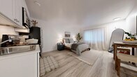 Located at 8835 S Hosmer St in Tacoma, WA, this efficiently designed one-bedroom, two-bathroom unit makes excellent use of its compact footprint across a single floor. The spacious primary bedroom serves as the central living area, featuring light wood-style flooring, crisp white walls, and a large window dressed with sheer curtains that allow abundant natural light to fill the room. A built-in kitchenette along one wall includes white shaker-style cabinetry, a stainless steel refrigerator, range, and microwave, providing all essential cooking amenities within easy reach. The room also accommodates a dining area with a rustic wooden table and stools, along with ample space for a full-sized bed and additional furnishings. The bathroom area is divided into two functional spaces: a shared vanity zone with a white shaker-style cabinet, a large mirror, and a textured accent wall, plus a separate water closet with light tile flooring and a full bathtub with a tiled surround. A hallway connects the spaces efficiently. The overall aesthetic is clean and contemporary, with a neutral palette of whites and light tones that enhance the sense of openness throughout.