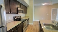 This single-story space showcases a well-appointed kitchen as its centerpiece, designed with both functionality and style in mind. The kitchen features rich, dark espresso-toned cabinetry that provides ample storage, complemented by light granite countertops that create an appealing contrast. Stainless steel appliances, including a smooth-top electric range, built-in microwave, and a stainless sink, lend the kitchen a contemporary, polished look.

The flooring throughout is a warm-toned light hardwood, lending continuity and a welcoming feel to the space. Neutral beige walls keep the atmosphere bright and versatile, while recessed ceiling-mounted lighting ensures consistent illumination throughout. The layout of the kitchen is efficiently designed in a galley-style configuration, maximizing the use of available space with countertops and cabinetry running along both sides.

A doorway visible at the end of the kitchen corridor leads into an adjacent room, suggesting an open and connected floor plan. Clean white baseboards and trim provide crisp finishing touches throughout, contributing to the overall tidy, move-in-ready condition of the space.