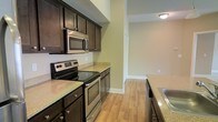 This single-story space showcases a well-appointed kitchen as its centerpiece, designed with both functionality and style in mind. The kitchen features rich, dark espresso-toned cabinetry that provides ample storage, complemented by light granite countertops that create an appealing contrast. Stainless steel appliances, including a smooth-top electric range, built-in microwave, and a stainless sink, lend the kitchen a contemporary, polished look.

The flooring throughout is a warm-toned light hardwood, lending continuity and a welcoming feel to the space. Neutral beige walls keep the atmosphere bright and versatile, while recessed ceiling-mounted lighting ensures consistent illumination throughout. The layout of the kitchen is efficiently designed in a galley-style configuration, maximizing the use of available space with countertops and cabinetry running along both sides.

A doorway visible at the end of the kitchen corridor leads into an adjacent room, suggesting an open and connected floor plan. Clean white baseboards and trim provide crisp finishing touches throughout, contributing to the overall tidy, move-in-ready condition of the space.