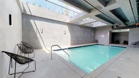 Located at 8201 Van Nuys Blvd in the Panorama City neighborhood of Los Angeles, this indoor pool facility presents a striking contemporary design defined by clean lines and a restrained material palette. The space is anchored by a generously sized rectangular swimming pool with vivid aqua-blue water, framed by cobalt blue tile trim along its perimeter and accessible via a stainless steel entry railing.

The architecture is characterized by smooth, poured concrete walls finished in a consistent cool gray tone, lending the space an urban, industrial-modern aesthetic. Large open ceiling cutouts and skylight apertures allow abundant natural light to filter in from above, casting dramatic angular shadows across the pool deck throughout the day and creating a dynamic interplay of light and shadow. Exposed overhead structural elements and utility conduits contribute to the raw, functional character of the design.

The surrounding pool deck offers ample circulation space, furnished with lightweight wire-frame lounge chairs and occasional accent seating positioned along the walls. A dedicated relaxation alcove with reclining chaise lounges provides a comfortable area for rest. Wall-mounted sconce lighting supplements the natural illumination. The overall ambiance is calm and contemplative, combining utilitarian architecture with refined minimalist detailing.