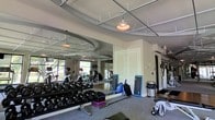 This single-floor fitness center offers a thoughtfully designed workout environment suited for a wide range of exercise routines. The space is anchored by a full wall of floor-to-ceiling mirrors that visually expand the room and allow users to monitor their form during training. A rack of dumbbells in varying weights lines the mirrored wall, accompanied by exercise mats and balance tools, providing a comprehensive free weights and functional training area. Cardio equipment, including stationary bikes and elliptical machines, is positioned toward the rear of the space, taking advantage of natural light streaming in through large windows that offer views of the surrounding greenery. The ceiling features a suspended grid design with decorative pendant lights that add a stylish touch to the otherwise functional environment. Additional equipment is accessible near the entrance, rounding out the facility's offerings. The flooring transitions from carpet in the stretching and free weight zone to a harder surface near the cardio and machine area, serving both comfort and durability needs. Overall, the space strikes a balance between practical fitness functionality and an inviting, well-lit atmosphere.