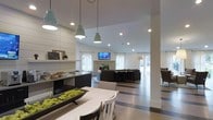 This single-floor community space offers a thoughtfully designed open-plan layout that seamlessly connects multiple functional zones, creating a cohesive and inviting environment. The interior is finished with a crisp white color palette accented by dark cabinetry and striped flooring, lending the space a clean, modern aesthetic.

The dining and lounge area at the front features a white table and chairs, complemented by striking teal pendant lights that hang at varying heights, adding a bold yet tasteful design element. Shiplap wall paneling and open shelving adorned with decorative accents, including a chalkboard sign, contribute a warm, farmhouse-inspired touch to the contemporary setting.

Recessed ceiling lighting throughout ensures the space is well-lit, while large windows and glass doors allow natural light to pour in, enhancing the open, airy feel. A wall-mounted flat-screen television anchors the main lounge area, which is furnished with dark upholstered seating arranged for comfortable gathering and relaxation. A fireplace serves as an architectural focal point within the lounge zone.

The layout flows effortlessly from one area to the next, supported by structural columns that define zones without interrupting the sense of spaciousness. Overall, the space delivers a polished, functional environment well-suited for socializing and community engagement.