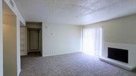 This single-floor residential space presents a straightforward and functional layout designed for comfortable everyday living. The main living area is a standout feature, offering a generous open floor plan finished with wall-to-wall plush carpeting that adds warmth and comfort underfoot. A large sliding glass door dressed with vertical blinds allows an abundance of natural light to flood the room, creating a bright and airy atmosphere. The neutral, cream-toned walls provide a versatile backdrop suitable for a variety of interior styles. The textured popcorn ceiling adds a classic touch characteristic of the space's construction era. A hallway visible from the living area leads to additional rooms, hinting at a practical and well-organized floor plan. Light switches and electrical outlets are thoughtfully placed throughout, ensuring convenience across the space. The overall ambiance is clean, open, and move-in ready, making it well-suited for those seeking a simple yet comfortable living environment. The single-floor design ensures easy accessibility and seamless navigation throughout the entire space.