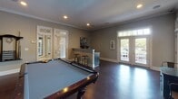 Located at 117 Abberly Green Blvd in Mooresville, NC, this single-floor clubroom is a well-appointed recreational space designed for community gathering and entertainment. Rich, dark hardwood flooring runs throughout, anchoring the room's sophisticated yet relaxed atmosphere. Crown molding and recessed lighting add refined architectural detail to the neutral gray walls.

At the heart of the space is a full-size billiards table, offering a central focal point for recreation. A built-in wet bar with granite countertops, an undermount sink, navy cabinetry, and bar stool seating provides a convenient serving area ideal for entertaining. Adjacent to the bar, a coffee and beverage station adds everyday convenience.

Multiple seating vignettes are arranged throughout the room, including industrial-style high-top tables with metal-and-wood bar chairs. Wall-mounted flat-screen televisions and curated abstract artwork enhance the social ambiance. Natural light flows in generously through several sets of glass-paneled French doors on multiple walls, offering outdoor access and a bright, open feel. The overall design balances comfort and function, making this a versatile space suited for both casual gatherings and planned community events.