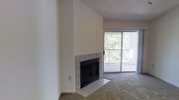 This single-floor space offers a clean, functional layout with a welcoming living area as its centerpiece. The room is anchored by a classic tile-surround fireplace, featuring a black firebox set against crisp white walls that rise to a textured ceiling, creating a cozy and timeless focal point. The flooring combines soft, wall-to-wall carpeting in the main living area with tile accents framing the fireplace hearth, providing both comfort and durability.

Natural light fills the space through a large sliding glass door, which opens onto a private balcony enclosed by a metal railing. The balcony overlooks a lush backdrop of mature trees, offering a peaceful, nature-filled view that brings a sense of the outdoors inside. The sliding door allows for easy indoor-outdoor flow and contributes to the bright, airy atmosphere of the living area.

The white walls throughout the space provide a neutral canvas that allows for versatile decorating possibilities. The textured ceiling and understated finishes give the space a practical and livable character. Overall, this space combines essential comforts — a fireplace, natural light, outdoor access, and greenery views — into a straightforward and inviting single-floor layout.