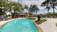 Located at 5500 De Soto St in Houston, Texas, this residential community's outdoor amenity area is designed to offer a resort-like experience within a well-maintained, landscaped setting. The centerpiece of the space is a generously sized freeform swimming pool with a striking natural stone surround and a decorative rock water feature, lending the area a relaxed, tropical ambiance. The pool's curvilinear shape and clear turquoise water create an inviting focal point, complemented by wide concrete pool decking that provides ample space for lounging.

The community buildings surrounding the pool area are constructed with brick and neutral-toned siding, featuring large windows, pitched rooflines, and covered stairwells. Mature trees, including towering pines and broad-canopy hardwoods, provide natural shade throughout the grounds, enhancing the outdoor experience. Thoughtfully placed landscaping with tropical-style plants and mulched garden beds adds color and texture to the environment.

A gated playground area is visible nearby, equipped with colorful play structures set on a grassy lawn, making the space family-friendly. Iron perimeter fencing encloses key areas for safety and privacy. Covered pergola structures offer additional shaded gathering spots, rounding out a comprehensive outdoor amenity space well-suited for recreation and relaxation.