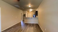 This single-floor residence showcases a thoughtfully designed open-concept layout that connects the living area and kitchen in a cohesive, functional flow. Rich, dark hardwood-style flooring runs throughout the main living space, adding warmth and a polished aesthetic that contrasts elegantly with the neutral, light-toned walls.

The kitchen is positioned centrally and features a peninsula-style breakfast bar with granite-look countertops and bar seating, making it an ideal spot for casual dining or entertaining. Dark cabinetry and black appliances provide a bold, modern contrast to the lighter surroundings, while recessed lighting and flush-mount ceiling fixtures ensure the space is well-lit throughout.

A ceiling fan in the living area adds both functionality and a finishing touch to the space. Overhead, HVAC vents are integrated cleanly into the ceiling, contributing to the tidy, uncluttered appearance of the home. A doorway visible in the background leads to additional areas of the residence, suggesting a practical and efficient floor plan. White interior doors and clean white baseboards complete the crisp, move-in-ready presentation of this well-maintained space.
