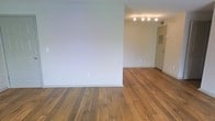 This single-floor space offers a straightforward and functional layout characterized by its warm, honey-toned hardwood floors that extend throughout the open living area. The floors display natural wood grain variation, adding texture and character to the otherwise clean aesthetic. Walls are finished in a crisp, neutral white, creating a bright and versatile backdrop suitable for a variety of interior styles. A partial wall divides the main living area from an adjoining space, offering a sense of definition without fully closing off the floor plan. In the adjacent area, a modern track lighting fixture is mounted to the ceiling, providing focused, adjustable illumination. A standard interior door leads further into the unit, and a wall-mounted panel — likely an electrical box or intercom — is visible nearby. Baseboard trim runs consistently along the walls, adding a clean finishing detail. The overall ambiance is minimal and move-in ready, with a neutral palette that allows for personalization. The space benefits from its open arrangement, making it practical and easy to configure for everyday living.