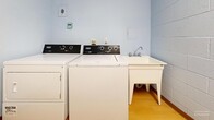 This compact laundry room is a single-floor utility space designed to serve residents with all essential washing amenities. The room is constructed with painted white and light blue cinder block walls, giving it a durable, no-frills character typical of shared-use facilities. The flooring throughout is a warm honey-toned hardwood-style surface that adds a touch of warmth to the otherwise utilitarian environment.

The laundry area is equipped with two Maytag top-loading washing machines and at least one dryer, providing ample capacity for regular use. A freestanding white utility sink with a gooseneck faucet is positioned nearby, offering a convenient handwashing or pre-treatment station. Built-in mint-green laminate shelving is mounted along one wall, providing storage space for laundry supplies and personal items.

A baseboard heater along one wall ensures the room remains comfortable year-round. Electrical outlets and a light switch are accessible near the entry door, which features a classic lever-style handle. Notices posted on the walls indicate that the laundry room is reserved for residents only, reinforcing its role as a dedicated communal amenity. The overall layout is practical and efficiently organized to make the most of the available space.