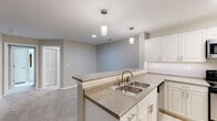 Located at 3301 Glenn McConnell Pkwy in Charleston, South Carolina, this single-floor residence offers a thoughtfully designed layout with clean, neutral finishes throughout. The kitchen is a functional centerpiece, featuring white cabinetry, granite-look laminate countertops, a double stainless steel sink set into a peninsula with pendant lighting overhead, and stainless steel appliances including an electric range with over-the-range microwave and a dishwasher. Tile flooring runs through the kitchen and utility areas, while plush carpeting extends across the living areas and bedrooms. The open living area is anchored by a low half-wall partition and features French doors that invite natural light and provide outdoor access. Bedrooms are comfortably sized with large windows overlooking greenery, ceiling fans with lighting, and soft carpeted floors. Two full bathrooms serve the unit — one featuring a tiled tub-shower combo with white vanity, and another with a frameless sliding glass shower door, white cabinetry, and tile flooring. A dedicated laundry room with wire shelving, tile flooring, and an electrical panel provides practical utility space. Textured ceilings and soft gray and beige wall tones create a cohesive, understated aesthetic throughout.