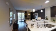 This well-appointed single-floor residence showcases a thoughtfully designed open-concept layout that seamlessly connects the kitchen, dining, and living areas. The kitchen features a sleek white breakfast bar illuminated by elegant pendant lights, creating a bright and functional centerpiece for the space. Rich dark hardwood flooring flows throughout, providing a warm contrast to the contemporary finishes. The dining area is defined by a bold dark accent wall, adding depth and sophistication to the overall aesthetic. The living room is generously proportioned, offering ample space for relaxation and entertaining. Large sliding glass doors flood the interior with natural light and open directly onto a private balcony, where lush greenery and outdoor seating can be enjoyed. The carefully curated color palette of neutral tones, accented by yellow and black textiles, lends the space a cohesive and modern character. Recessed lighting and stylish floor lamps complement the pendant fixtures, ensuring the space is well-lit throughout. The overall ambiance is one of contemporary comfort — polished, inviting, and designed with both function and aesthetics in mind.
