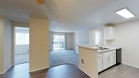 This single-floor residence offers an open and functional layout designed to maximize flow between living spaces. The kitchen is positioned at the heart of the home, featuring a peninsula with a built-in sink, white cabinetry providing ample storage, and light countertops. The kitchen opens directly into the main living area, making it ideal for everyday living and entertaining. The living room is generously sized, finished with plush blue-grey carpeting that adds warmth and comfort underfoot. A sliding glass door fills the room with natural light and provides direct access to a private outdoor balcony, creating a seamless connection between indoor and outdoor living. The entryway and kitchen area feature rich dark hardwood flooring, offering a stylish contrast to the carpeted living space. Textured ceilings are present throughout, and the warm yellow accent wall in the transition area adds a touch of color to the otherwise neutral palette of whites and soft greys. A ceiling fan is visible near the entry, contributing to the home's practical amenities. Overall, the space presents a clean, bright, and welcoming atmosphere well-suited for comfortable daily living.