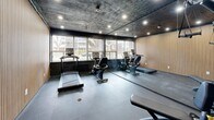 Located at 1 Marquette Ave in Toronto's vibrant west end, this single-room fitness facility offers a well-appointed workout space within a compact footprint. The room is designed with a bold, contemporary aesthetic, featuring a striking dark-painted ceiling paired with recessed pot lighting that creates a focused, energetic atmosphere. One wall is elegantly clad in warm, vertically slatted wood paneling, providing a rich textural contrast against the darker surroundings. The flooring is a durable rubberized surface suited for exercise use, while the walls incorporate large sliding windows that invite ample natural light and offer views of the surrounding greenery and neighbourhood streetscape. The space is equipped with cardio machines including treadmills and a stationary bike, as well as a multi-function strength training station, making it a well-rounded facility for a variety of workout routines. A full-length mirror along one wall visually expands the room and supports form-checking during exercise. A wall-mounted mini-split unit ensures comfortable climate control year-round. The entry features a dark metal door, reinforcing the space's modern, utilitarian design sensibility. Overall, this fitness room delivers a sleek and functional environment well-suited for residents seeking convenient, on-site exercise amenities.