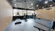 Located at 1 Marquette Ave in Toronto's vibrant west end, this single-room fitness facility offers a well-appointed workout space within a compact footprint. The room is designed with a bold, contemporary aesthetic, featuring a striking dark-painted ceiling paired with recessed pot lighting that creates a focused, energetic atmosphere. One wall is elegantly clad in warm, vertically slatted wood paneling, providing a rich textural contrast against the darker surroundings. The flooring is a durable rubberized surface suited for exercise use, while the walls incorporate large sliding windows that invite ample natural light and offer views of the surrounding greenery and neighbourhood streetscape. The space is equipped with cardio machines including treadmills and a stationary bike, as well as a multi-function strength training station, making it a well-rounded facility for a variety of workout routines. A full-length mirror along one wall visually expands the room and supports form-checking during exercise. A wall-mounted mini-split unit ensures comfortable climate control year-round. The entry features a dark metal door, reinforcing the space's modern, utilitarian design sensibility. Overall, this fitness room delivers a sleek and functional environment well-suited for residents seeking convenient, on-site exercise amenities.
