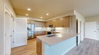 This single-story home features a thoughtfully designed layout accommodating four bedrooms and two full bathrooms. The open-concept kitchen and living area serves as the heart of the home, showcasing modern flat-front cabinetry in a warm wood-grain finish, light quartz countertops, a subway tile backsplash, stainless steel appliances including a French-door refrigerator, and a central island with an undermount sink. An adjacent pantry provides additional storage. The living room receives ample natural light through large windows and a glass exterior door that opens to an outdoor area, offering views of the surrounding landscape. Light hardwood-style flooring runs throughout the main living spaces, while bedrooms feature plush carpeting. Each bedroom includes closet storage, with one bedroom featuring a walk-in closet equipped with adjustable shelving and hanging rods. Secondary bedrooms include sliding-door closets with built-in shelving. Both bathrooms are finished with wood-tone vanity cabinetry, white quartz countertops, and a shower-tub combination. A dedicated laundry room houses a full-size washer and dryer. The overall aesthetic is clean and contemporary throughout.