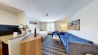 Located at 20 International Place in Tewksbury, MA, this efficiently designed suite offers a comfortable and functional layout across a single floor. The open-concept interior is anchored by a carpeted living area featuring a blue sofa and armchair ensemble, a coffee table, and framed wall art — all illuminated by natural light from a large window with sheer curtains. A built-in workspace with track lighting lines the entryway corridor, making this suite well-suited for extended stays or remote work. The bedroom area is seamlessly integrated into the main space, featuring a queen bed, dresser with flat-screen TV, and direct sightlines to the compact kitchen. The kitchen includes dark cabinetry, granite-style countertops, and essential appliances. The bathroom is clean and bright, finished with wood-look flooring, a granite-topped vanity with dark wood cabinetry, and a full tub/shower combination with a curtain enclosure. A closet provides additional storage. The neutral color palette, patterned carpet, and consistent modern finishes give the suite a cohesive, polished appearance throughout.