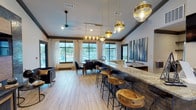 Located at 6721 Washington Ave in Ocean Springs, Mississippi, this single-floor clubhouse facility combines sophisticated design with inviting communal spaces. The interior features light wood-look tile flooring, dark crown molding, and vaulted ceilings that lend an open, airy feel throughout.

A standout centerpiece is the large bar counter clad in textured stone with a striking marble-look countertop, lined with rustic live-edge wooden barstools and illuminated by a series of gold-accented globe pendant lights. Adjacent to the bar, a spacious dining area accommodates a large wooden table surrounded by dark leather chairs, with floor-to-ceiling windows framed by sheer white curtains offering direct views of the outdoor pool area.

The lounge area features a plush cream sofa and navy accent chairs arranged around a stylish coffee table, anchored by a richly textured wood-paneled feature wall near the entrance. Natural light floods this area through oversized windows, creating a warm and welcoming atmosphere.

A semi-private office or media nook is set apart by glass-paneled doors and showcases a dramatic tile and wood accent wall with a wall-mounted television, accompanied by a sleek upholstered chair — ideal for focused work or quiet retreat. The cohesive mix of gold hardware, warm wood tones, and stone textures gives this clubhouse a refined yet comfortable character.