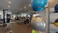 This fitness center is a comprehensively outfitted single-floor facility designed to support a wide range of workout styles and fitness goals. The space is finished with durable rubber flooring throughout and features recessed lighting that keeps the environment well-lit and energetic. Floor-to-ceiling mirrors line one full wall, visually expanding the room while allowing users to monitor their form during exercise.

The cardio zone includes a row of treadmills and elliptical machines positioned along the mirrored wall, providing an open and motivating workout environment. Natural light filters in through several windows fitted with blinds, complementing the interior lighting. A dedicated strength training area houses a cable pulley machine, weight benches, and a punching bag station complete with speed bag attachment.

The free weights section is thoroughly stocked with a full rack of dumbbells and kettlebells spanning a wide range of weights, alongside a TAG-branded rack holding a complete set of medicine balls. Stability and exercise balls in multiple sizes are neatly organized on a dedicated rack. Large glass panel windows along one wall provide a visual connection to an adjacent lounge or common area, adding a sense of openness. Direct exterior door access adds convenient entry and exit for users.
