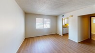 This single-floor space features a straightforward, functional layout with consistent design elements carried throughout. The living area is bright and open, benefiting from a window that allows natural light to fill the room and illuminate the warm wood-style laminate flooring underfoot. A textured popcorn ceiling adds a classic finish overhead, while crisp white walls maintain a clean, neutral atmosphere. A floor vent and wall outlets are conveniently positioned throughout the space.

An additional room, likely serving as a bedroom, mirrors similar finishes with matching laminate flooring, white walls, and a textured ceiling. A ceiling fan with an integrated light fixture provides both air circulation and overhead lighting, while a window fitted with blinds allows for adjustable natural light and privacy.

The galley-style kitchen is efficiently arranged, featuring white cabinetry, a double-basin sink, a gas range, and a refrigerator. Laminate countertops provide practical workspace, and a small window above the sink brings in additional light. The kitchen connects directly to the living area, supporting an easy flow between spaces.

The bathroom is neatly appointed with tile flooring, a white vanity with cabinet storage, a toilet, and a shower-tub combination with tile surround and a curtain rod. A window above the tub provides ventilation and natural light.