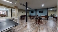 Located at 300 Canal in Lawrence, Massachusetts, this single-floor amenity space serves as a dynamic recreational hub designed for social engagement and leisure. The main game room features warm, wood-look plank flooring throughout, complemented by bold black structural columns that add an industrial character to the space. Multiple billiards tables take center stage, alongside a ping-pong table, foosball table, and a shuffleboard table, offering a wide variety of entertainment options. High bar-height tables with padded stools are strategically placed throughout the room, providing comfortable spots for socializing or watching one of the wall-mounted flat-screen televisions. The space is illuminated by linear overhead lighting that ensures a well-lit environment. A standout architectural feature is the curved glass partition wall with sleek full-length glass panels and polished chrome hardware, elegantly separating areas while maintaining visual openness. Through the glass, a connected dining or lounge area is visible, featuring pendant lighting and brick-accented walls, hinting at the broader community amenities available. The flooring transitions to large-format stone-look tile within the glass-enclosed section, adding a refined contrast to the rustic wood of the main game room. Overall, the space strikes a balance between industrial and modern design elements.