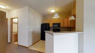 This single-floor residence offers a functional and well-organized layout designed for comfortable everyday living. The kitchen serves as a central highlight, featuring warm honey-toned wood cabinetry that provides ample storage space. Black appliances, including a refrigerator, microwave, and range, lend a clean, consistent look to the cooking area. A peninsula with a light-toned countertop extends the workspace and creates a natural division between the kitchen and the adjacent living space, making it ideal for casual dining or meal preparation. Pendant lighting hangs above the peninsula, adding focused illumination and a touch of character to the space. The flooring transitions from tile in the kitchen area to carpet in the living zone, subtly defining each functional space. Neutral-toned walls throughout keep the atmosphere light and versatile, providing a blank canvas for personalization. Flush ceiling lighting ensures the interior remains well-lit. The overall design emphasizes practicality and ease of movement, with an open flow that connects the kitchen and living areas into a cohesive, inviting environment suited for a range of lifestyles.