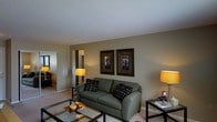 This single-floor space features a welcoming living area centered around a plush, sage-green sofa accented with patterned throw pillows. A glass-topped coffee table sits in front of the sofa, adding a contemporary touch to the room. Two framed artworks in dark ornate frames adorn the neutral-toned walls, contributing a refined aesthetic. Warm ambient lighting is provided by table lamps on either side of the seating area, creating an inviting atmosphere. The room features light-colored carpeting that complements the earthy wall tones. An open doorway visible from the living area provides a glimpse into an adjoining room, suggesting a connected and functional layout. The textured ceiling adds subtle architectural character. Overall, the space maintains a balanced, comfortable ambiance with a neutral color palette, practical furnishings, and thoughtful decor that creates a lived-in yet organized environment suitable for relaxing or entertaining.