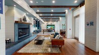 This thoughtfully designed single-floor clubhouse combines modern farmhouse aesthetics with upscale amenities throughout its open, flowing layout. Exposed dark wood ceiling beams contrast beautifully against a muted teal ceiling and shiplap-clad walls, creating a warm yet contemporary ambiance. Rich hardwood flooring extends throughout the entire space, anchoring each distinct area with warmth and continuity.

The main lounge area serves as the heart of the space, featuring a striking fireplace, curated seating arrangements with layered area rugs, and a large-format video display wall ideal for entertainment. Adjacent to the lounge, floor-to-ceiling windows flood the space with natural light and offer direct views of the outdoor courtyard, with cozy nook seating positioned to take full advantage of the scenery.

A well-appointed wet bar area stands out with bold teal cabinetry, marble countertops, a farmhouse-style stainless steel sink with brass bridge faucet, white subway tile backsplash, and open wood shelving — a refined and functional gathering spot. A game room tucked nearby features a shuffleboard table beneath a decorative pressed-tin ceiling and patterned wallcovering, adding a playful, eclectic character. Steel-framed glass partitions and a multi-screen display wall complete this dynamic, amenity-rich environment.