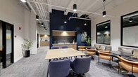 Located at 628 Ellicott St in Buffalo, NY, this single-floor professional workspace delivers a well-designed blend of collaborative and private environments. The open central area anchors the suite with a large light wood communal table surrounded by upholstered navy blue rolling chairs, set atop textured gray carpet. A dramatic navy accent wall frames a built-in kitchenette equipped with wood cabinetry, a tile backsplash, sink, microwave, and coffee station — ideal for daily workplace needs. Adjacent to the main workspace, a comfortable lounge area features warm leather seating and a side table, providing a relaxed setting for informal meetings or breaks. Exposed white structural ceilings with pendant lighting and visible ductwork lend an industrial-modern character to the space, while large black-framed windows invite natural light throughout. A dedicated formal conference room offers a long wood table with ample seating, recessed lighting, and locally inspired artwork on the walls, creating a polished setting for client meetings. Private phone booths and glass-partitioned focus rooms along the corridor offer quiet, enclosed spaces for individual work or calls, rounding out a thoughtfully designed office environment suited for productivity at every level.