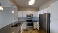 This single-floor residence offers a thoughtfully designed open-concept layout that connects the living and kitchen areas seamlessly. The kitchen is a functional and stylish centerpiece, featuring white shaker-style cabinetry, dark countertops, and a full suite of stainless steel appliances including a refrigerator, electric range, and over-the-range microwave. A double-basin sink with a modern faucet and pendant lighting overhead add refined finishing touches. The kitchen flows naturally into the living area, which is finished with plush neutral-toned carpeting that adds warmth underfoot. A built-in niche with white cabinetry provides a convenient storage solution and a subtle architectural detail along one wall. The space is painted throughout in warm greige tones that lend a calm, cohesive ambiance. Ceiling-mounted vents ensure consistent climate control, and a wall-mounted thermostat offers easy access. Sconce lighting along the walls contributes to the overall warm and welcoming atmosphere. The layout is efficient and well-organized, making excellent use of the available footprint while maintaining an open and airy feel throughout.