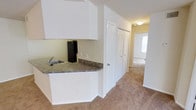 This single-floor residence offers a practical and straightforward layout designed for comfortable everyday living. The kitchen is positioned at the front of the unit and features white upper cabinetry, dark granite-style laminate countertops, a stainless steel sink, and a black refrigerator. A partial countertop peninsula separates the kitchen from the adjoining carpeted hallway, creating a subtle open-concept feel while maintaining defined functional zones.

The hallway extends from the kitchen area toward the rear of the unit, lined with neutral beige walls and soft carpeted flooring that continues throughout the living spaces. A louvered bifold closet door along the hallway provides convenient storage access. The textured ceiling is consistent throughout, adding a classic residential character to the interior.

At the end of the hallway, an additional room is visible, benefiting from natural light entering through a window. The overall color palette is light and neutral, dominated by whites and warm beiges, creating a clean and understated atmosphere throughout the space.
