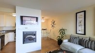 This single-floor space presents a cohesive, modern interior centered around a striking white-paneled feature wall that houses both a wall-mounted television and a built-in fireplace below it, creating a clean and functional focal point in the living area. The wood-look flooring runs throughout, adding warmth and continuity between the living space and an adjacent dining or breakfast nook area visible in the background. That secondary area is furnished with a bistro-style high table and barstool seating beneath a ceiling fan with integrated lighting, offering a casual spot for dining or casual gatherings. The living area itself is accented by a starburst wall clock or mirror, a lush potted floor plant, and a light gray sofa with patterned throw pillows, contributing to a relaxed yet stylish atmosphere. The walls are finished in a soft, neutral tone that keeps the space feeling bright and open. The overall layout is efficient and well-organized, with a clear visual connection between the living and dining zones. Decorative touches throughout give the space personality while maintaining a clean, uncluttered look suitable for comfortable everyday living.