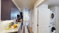 This single-floor unit offers a thoughtfully designed layout that maximizes functionality across its key living spaces. The kitchen runs along one wall in a galley-style configuration, finished with dark cabinetry, light quartz countertops, a subway tile backsplash, and a full suite of stainless steel appliances including an electric range, over-the-range microwave, and refrigerator. Light hardwood flooring extends throughout the unit, creating a cohesive and warm aesthetic from room to room.

The bathroom is generously sized and serves a dual purpose, housing a stacked washer and dryer unit alongside the primary bath fixtures. A wide vanity with dual sinks, dark wood cabinetry, and quartz countertops provides ample storage and prep space. A tub-shower combination with a patterned curtain completes the bath area.

The living space is open and bright, benefiting from multiple windows that invite natural light. A glass-paneled exterior door leads to an outdoor area, further enhancing the airy feel of the room. A sliding barn-style door adds a modern architectural touch, separating spaces with both style and practicality. Overall, the unit presents a clean, contemporary aesthetic.