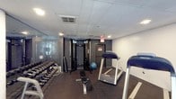 This single-level fitness space is designed to accommodate a variety of workout routines within a efficiently organized layout. The room features a dropped ceiling with recessed ventilation and standard lighting, contributing to a functional, no-frills gym atmosphere. Dark rubberized flooring covers the workout area, providing both safety and durability underfoot.

Along one wall, a mirrored panel extends the visual depth of the space while allowing users to monitor their form during exercise. A multi-station cable machine serves as a centerpiece for strength training, flanked by a flat workout bench ideal for a range of exercises. A stability ball adds versatility to the available equipment.

A well-stocked dumbbell rack lines one side of the room, holding an organized selection of free weights in varying sizes, catering to both beginner and advanced users. On the cardio side, at least two treadmills are positioned to provide ample space for users during peak hours. An illuminated emergency exit sign and clearly posted safety notices reflect adherence to building safety standards. Overall, the space delivers a practical and comprehensive fitness environment.