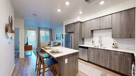This contemporary single-floor residence is thoughtfully designed with an open-concept layout that seamlessly connects the kitchen and living areas. The kitchen is a standout feature, showcasing wood-grain cabinetry in a warm gray tone, a classic white subway tile backsplash, and a suite of stainless steel appliances including a refrigerator, dishwasher, and range with microwave. A central island with a crisp white countertop provides seating for multiple bar stools, making it an ideal space for casual dining and entertaining. The living area benefits from generous natural light streaming through large windows and a glass-panel door, which also offers access to the outdoors. Light wood-style flooring flows consistently throughout the unit, creating a cohesive and airy feel. The bedroom is a calm retreat, featuring neutral tones, clean lines, and ample room for furnishings. The bathroom includes a soaking tub and shower combination with large-format tile surround and wood-look flooring. A practical dedicated laundry area houses a stacked washer and dryer unit alongside built-in shelving for added storage. A clean entryway with multiple closet doors rounds out this well-organized and functional living space.