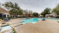 Located at 2300 Kathryn Ln in Plano, Texas, this two-story residential community offers a well-appointed outdoor amenity area that serves as a central gathering space for residents. The centerpiece of the outdoor environment is a generously sized swimming pool with clearly marked lap lanes, providing both recreational and fitness-oriented use. Adjacent to the main pool sits a stone-finished spa with a waterfall feature, surrounded by decorative white river rock and ornamental grasses, adding a resort-like aesthetic to the setting.

The pool deck is finished in a textured aggregate surface and is furnished with rows of blue lounge chairs and seating arrangements, offering ample space for relaxation. A covered pergola with bar-height seating is positioned along the perimeter, providing a shaded outdoor dining and social area. Mature native trees are distributed throughout the grounds, offering natural shade and a sense of established, lush landscaping. Vibrant blue ceramic planters with evergreen topiaries add decorative accents along the walkways.

The surrounding two-story buildings feature pitched rooflines with architectural gable details and traditional exterior finishes, framing the outdoor space and contributing to the cohesive community aesthetic. The overall ambiance is that of a well-maintained, amenity-rich residential community designed for comfortable outdoor living.