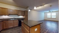 Located at 921 S Orlando Ave in Maitland, Florida, this single-floor residence offers a thoughtfully designed open-concept layout that seamlessly connects the kitchen and living areas. The kitchen serves as the focal point of the space, showcasing warm honey-toned cabinetry with detailed paneling, dark granite countertops, and a central island that provides additional workspace and storage. Stainless steel appliances, including a refrigerator and microwave, complement the black range and dishwasher, while pendant lighting and a track lighting system overhead add both function and visual interest. Crown molding accents the kitchen and living areas, lending a refined architectural touch. Rich, dark hardwood-style flooring flows consistently throughout the entire unit, creating a cohesive and grounded aesthetic. The bedroom areas are bright and airy, benefiting from natural light through large windows dressed with horizontal blinds. A ceiling fan in one bedroom adds comfort. Built-in shelving in the hallway offers practical storage solutions. The bathroom features matching wood cabinetry with granite countertops, a tiled tub-shower surround, and coordinating wood-look flooring. Overall, the space balances classic warmth with functional, modern design elements.