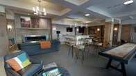 This single-floor community lounge space offers a thoughtfully designed, open-concept layout that balances comfort with a contemporary aesthetic. The room is anchored by a sleek, linear gas fireplace set against a stacked stone accent wall, paired with plush sofa seating and warm decorative throws — creating an inviting area for relaxation. Colorful, large-format artwork adds visual interest throughout the space.

At the heart of the room, a set of high-top bistro tables and industrial-style bar stools provide casual seating ideal for socializing or working. These flow naturally into a bar or kitchenette area featuring a light-colored countertop, dark cabinetry, and a series of elegant pendant lights that hang above, adding warmth and definition to the space.

The ceiling features bold cross-beam architectural detailing that adds depth and structure to the room, while a combination of recessed lighting, pendant fixtures, and a statement chandelier creates layered illumination throughout. A wall-mounted flat-screen television is positioned for easy viewing from multiple seating areas. The carpeted flooring in a muted tone ties the various zones together, contributing to the overall cohesive and welcoming ambiance of this well-appointed communal space.