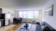 This single-floor space combines a living area and dining area in an open, functional layout. The living area is furnished with a spacious gray sectional sofa accented with patterned throw pillows, positioned around a sleek black coffee table atop a textured area rug. Adjacent to the living space, a dark wood dining table with matching chairs provides a dedicated area for meals and gatherings. A wall of large windows spans the length of the shared space, fitted with white roller shades that allow for adjustable natural light while offering views of greenery outside. The windows also house a built-in radiator and a wall-mounted air conditioning unit, ensuring year-round comfort. The flooring throughout is warm hardwood, adding a classic, inviting quality to the space. Walls are finished in a clean white, contributing to the bright and airy atmosphere. The textured ceiling adds subtle visual interest overhead. A floor lamp in the corner of the living area provides additional ambient lighting. Overall, the space presents a clean, modern aesthetic with practical furnishings suited to everyday living.