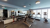 This modern fitness center is designed to accommodate a full spectrum of workout routines within a single, expansive floor. The space is finished with durable dark rubber flooring throughout, providing a professional athletic environment suited for both high-intensity and low-impact training.

The main workout area is lined with large floor-to-ceiling mirrors along one wall, visually expanding the space and allowing users to monitor their form. Circular halo-style light fixtures are mounted across the ceiling, providing consistent, ambient illumination. Natural light is introduced through windows along one end of the facility, enhancing the overall brightness of the space.

The cardio zone features multiple treadmills and a Life Fitness recumbent bike, offering a variety of aerobic training options. The strength training section is thoroughly outfitted with a full rack of dumbbells spanning a wide weight range, a medicine ball storage tower, stability balls, and multiple adjustable weight benches. A multi-functional cable machine and additional selectorized strength machines provide options for targeted resistance training.

A secondary alcove area houses additional selectorized machines, a narrow console table, and a Bluetooth speaker, creating a quieter zone for isolated exercises. The clean, neutral color palette of light gray walls paired with the dark flooring gives the facility a sleek, contemporary aesthetic throughout.