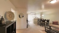 This single-floor space showcases an open-concept layout that effortlessly connects the living, dining, and kitchen areas into one cohesive and functional environment. The living area features plush wall-to-wall carpeting in a neutral tone, contributing to a warm and welcoming atmosphere throughout. A decorative round mirror and dark-finished accent furniture near the entryway add character and a sense of style upon arrival. The dining area is defined by a dark wood table and chairs set beneath a stylish pendant light, creating an inviting space for meals and gatherings. Flowing seamlessly from the dining area, the kitchen is outfitted with white cabinetry, a central island or peninsula, and a recessed ceiling light fixture that provides ample illumination for cooking and prep work. The consistent use of light, neutral tones on the walls and ceilings — including a textured ceiling finish — contributes to a bright, airy feel across the entire space. The open sightlines between rooms enhance the sense of spaciousness and make the layout particularly practical for everyday living and entertaining alike.