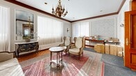 Located at 4555 Avenue Bonavista in Montréal, this distinguished space exudes timeless elegance across a single floor. The grand entrance lobby immediately sets a refined tone, showcasing floor-to-ceiling rich mahogany wood paneling, decorative raised moldings, and warm wall sconces that cast a welcoming glow. A stunning multi-arm brass chandelier anchors the ceiling, while a classical white marble statue greets visitors near the entry doors. Light travertine flooring and a central runner guide guests toward a staircase leading deeper into the building. The living room is a study in classical European design, featuring warm parquet hardwood floors in a herringbone pattern, intricate crown moldings, and delicate damask-patterned wallpaper set within arched panel frames. Large windows dressed with sheer white drapes allow generous natural light to fill the room. A second ornate brass chandelier echoes the grandeur of the lobby, complemented by brass wall sconces. An adjacent area extends the living space further, offering additional room with the same refined architectural detailing. Together, the spaces convey a sense of gracious, formal elegance with consistent classical architectural character throughout.