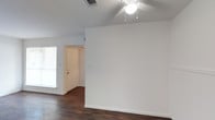 This single-floor residential space presents a straightforward and functional layout designed for comfortable everyday living. The main living area is characterized by crisp white walls that create a bright, neutral backdrop, complemented by rich, dark hardwood flooring that runs throughout and adds a sense of warmth and contrast to the space. A ceiling fan with a built-in light fixture is centrally mounted, providing both air circulation and overhead lighting to keep the room comfortable year-round. A window fitted with blinds allows natural light to filter into the space while offering privacy control. The entryway is accessible through a classic white interior door, providing a clear transition point into the home. Visible HVAC vents integrated into the ceiling contribute to the space's climate control infrastructure. The clean, uncluttered walls and open floor plan give the space a versatile quality, making it well-suited for a variety of furniture arrangements and interior styles. The overall ambiance is simple and move-in ready, with a neutral palette that serves as an adaptable canvas for personalization.